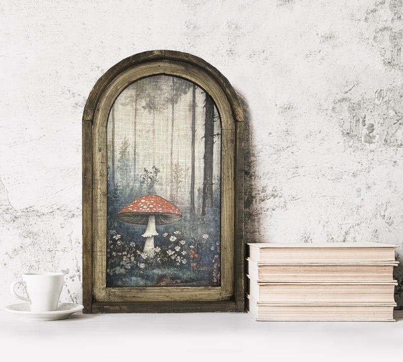 May include: A framed art print featuring a forest scene with a large red and white mushroom. The artwork is set in a rustic, arched wooden frame. A white teacup and saucer are in the foreground, with a stack of books to the right.