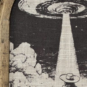 Puede incluir: Una ilustraci&oacute;n en blanco y negro de un OVNI sobrevolando un paisaje des&eacute;rtico con un vaquero o una vaquera montando a caballo en primer plano. El OVNI emite un rayo de luz.