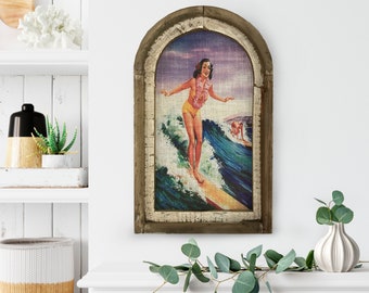 May include: Vintage-style framed artwork featuring a surfer girl riding a wave. The artwork has a distressed, arched wooden frame. The surfer is wearing a yellow swimsuit and a flower lei. The background shows a blue ocean and a purple sky.
