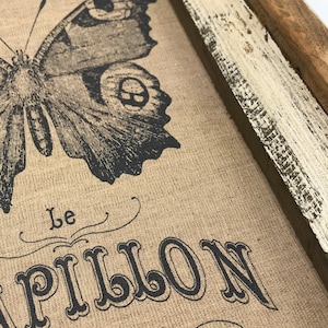 May include: A framed burlap print featuring a black and white butterfly illustration. The text "Le Papillon (The Butterfly)" is printed below the butterfly.