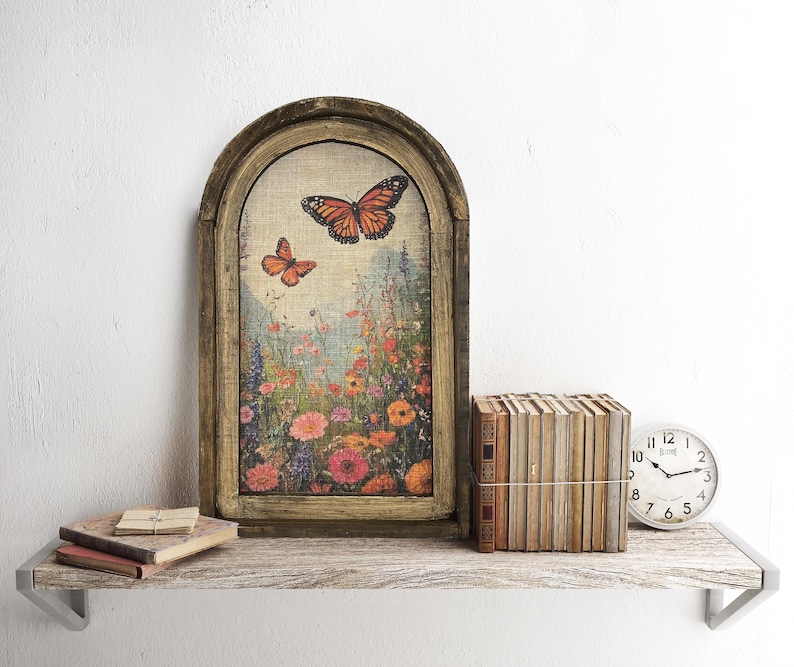 May include: An arched framed artwork featuring a butterfly and floral scene. The artwork is displayed on a rustic wooden shelf with a stack of books, a vintage clock, and small books with a clip.