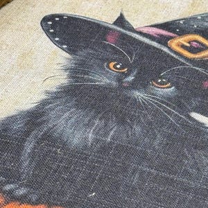 May include: A close-up of a black cat wearing a witch hat with a pink band and gold buckle. The cat has orange eyes and white whiskers. The image has a Halloween theme.