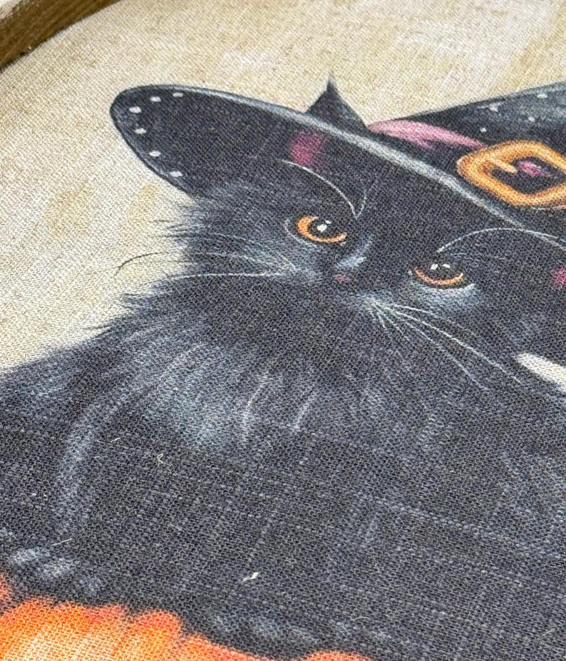May include: A close-up of a black cat wearing a witch hat with a pink band and gold buckle. The cat has orange eyes and white whiskers. The image has a Halloween theme.