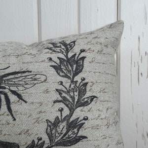 May include: A beige throw pillow with a black and white floral design featuring a bee and leaves. The pillow has a textured fabric.