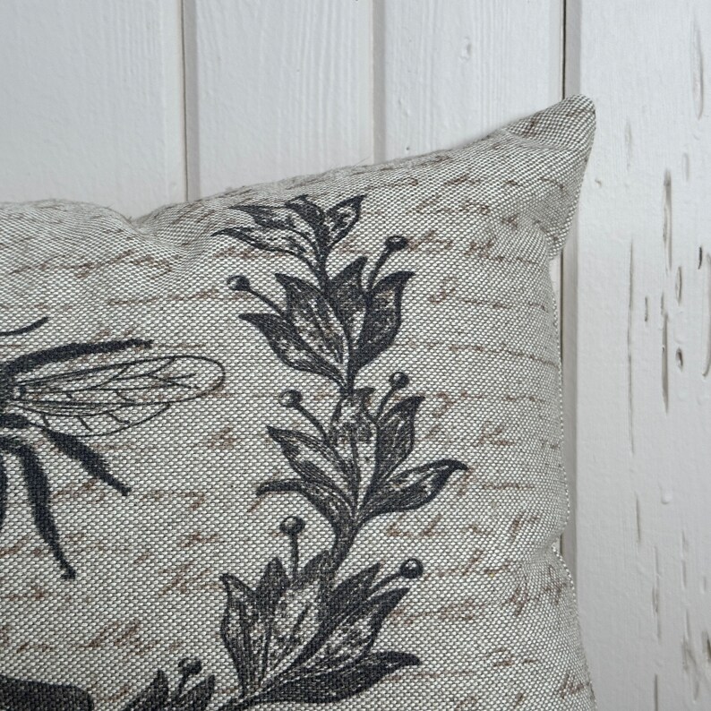May include: A beige throw pillow with a black and white floral design featuring a bee and leaves. The pillow has a textured fabric.