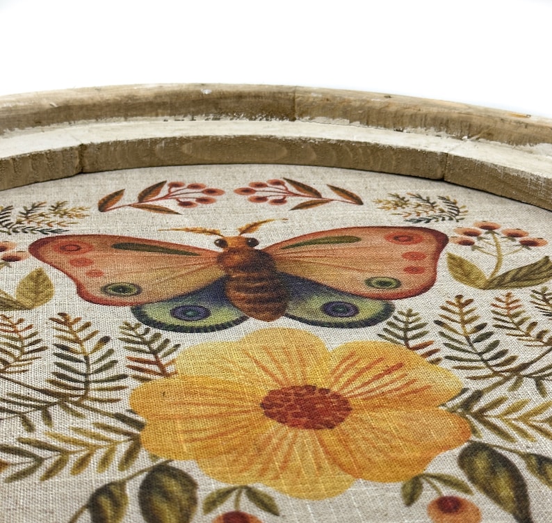 May include: A round wooden tray with a linen fabric insert featuring a floral design with a butterfly and yellow flower. The tray is perfect for serving snacks or drinks.