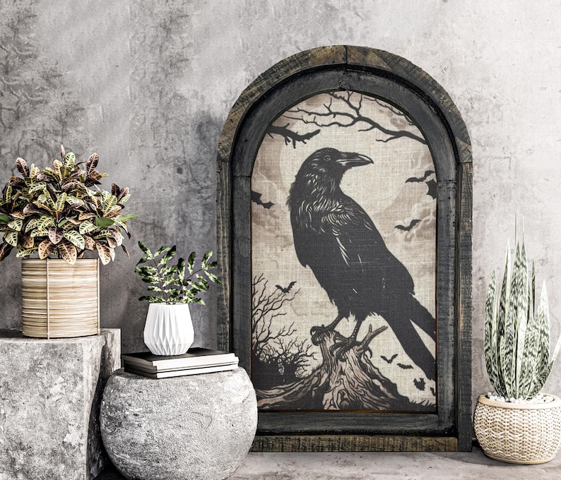 May include: A black raven silhouette on a beige canvas with a dark wood frame. The raven is perched on a tree branch with a dark background. The canvas is surrounded by a dark wood frame with a distressed finish. The image is displayed on a gray wall with two potted plants.