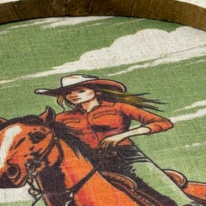 May include: A woman in a red shirt and cowboy hat rides a brown horse against a green and white background.