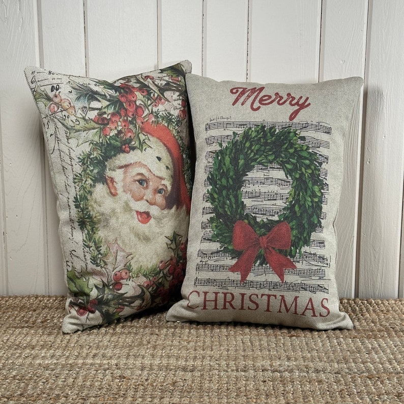 May include: Two decorative throw pillows with a Christmas theme. One pillow features a vintage illustration of Santa Claus with holly and berries. The other pillow has a wreath with a red bow and the words "Merry Christmas" on a musical note background.