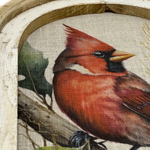 May include: A framed canvas print of a red cardinal perched on a branch with green leaves. The frame is a rustic wood with a whitewash finish.