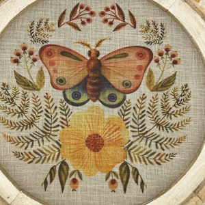 May include: A round framed wall art piece with a beige linen background featuring a stylized butterfly with orange and pink wings, a yellow flower with orange center, and green foliage.