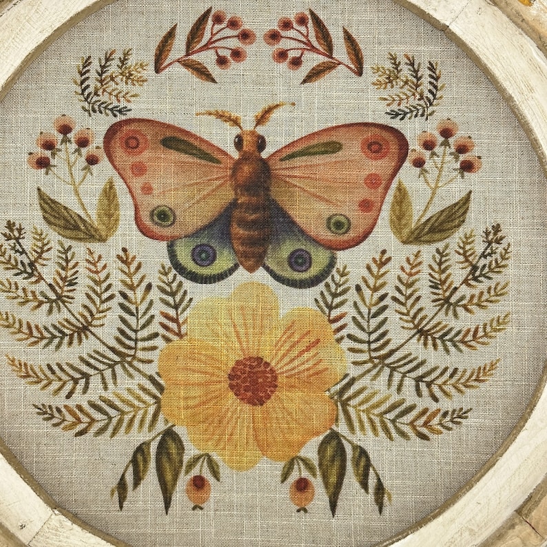 May include: A round framed wall art piece with a beige linen background featuring a stylized butterfly with orange and pink wings, a yellow flower with orange center, and green foliage.