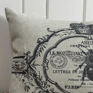 May include: A beige throw pillow with a black and white design featuring a bee, flowers, and the text "LE MOY" and "LETTRES ET JOURNAUX".