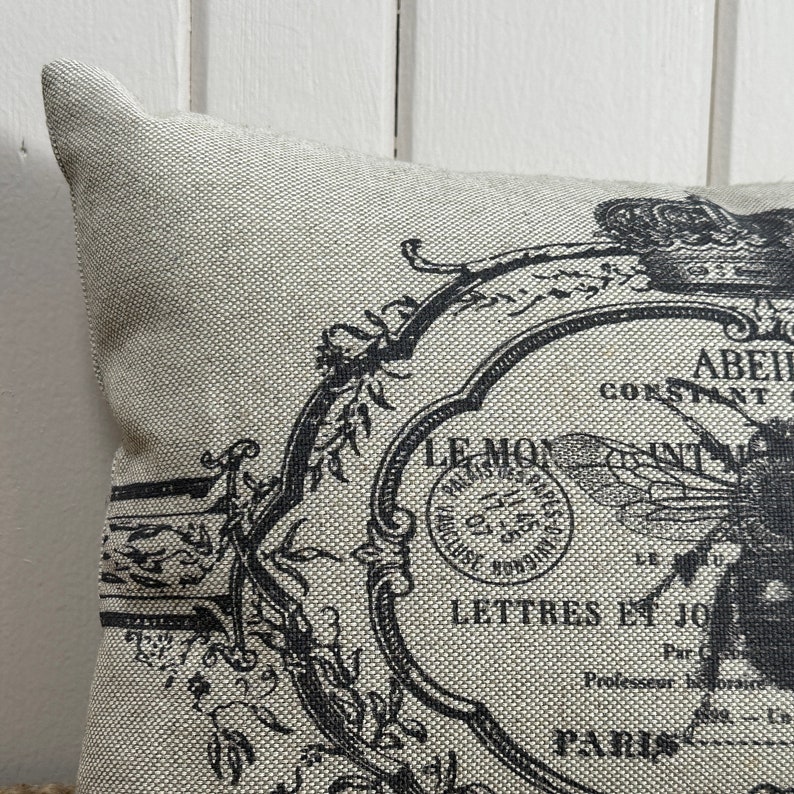 May include: A beige throw pillow with a black and white design featuring a bee, flowers, and the text "LE MOY" and "LETTRES ET JOURNAUX".