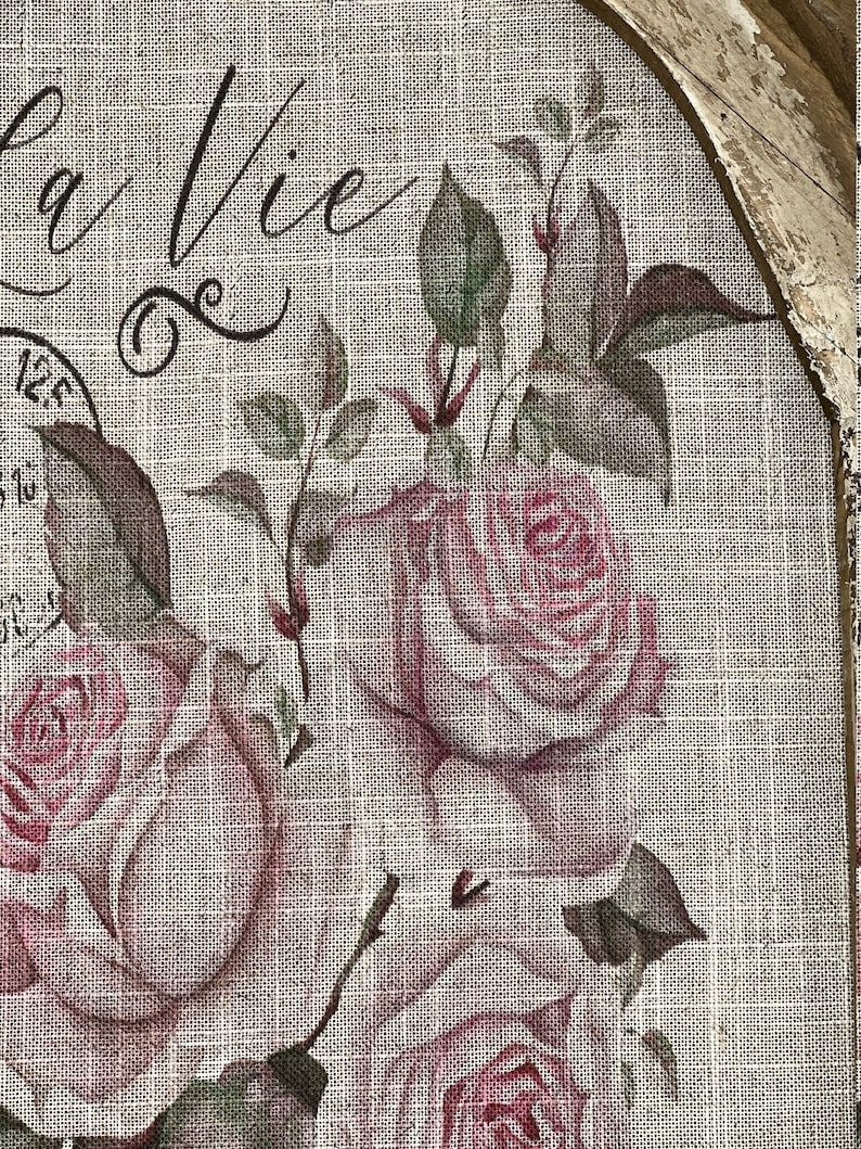 Pu&ograve; includere: Un primo piano di un tessuto di juta con un motivo floreale. Il tessuto presenta rose rosa e foglie verdi. Il testo "La Vie" &egrave; scritto in corsivo sul tessuto.