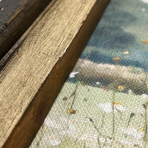 May include: Close-up of a framed landscape painting. The frame is made of dark brown and gold-painted wood. The painting depicts a field of white daisies and a distant hill under a blue sky.