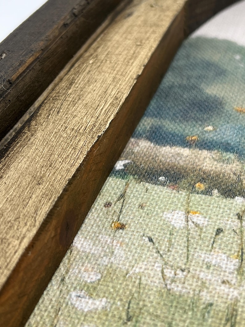 May include: Close-up of a framed landscape painting. The frame is made of dark brown and gold-painted wood. The painting depicts a field of white daisies and a distant hill under a blue sky.