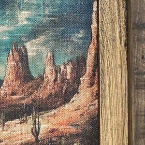 May include: A framed canvas print of a desert landscape with red rock formations, cacti, and a blue sky with clouds. The frame is made of wood with a rustic finish.
