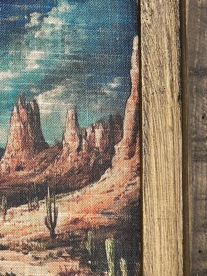 May include: A framed canvas print of a desert landscape with red rock formations, cacti, and a blue sky with clouds. The frame is made of wood with a rustic finish.