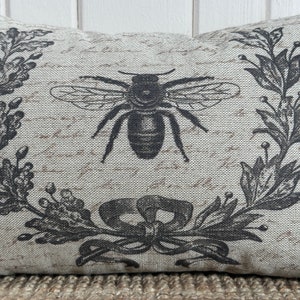 May include: A decorative pillow with a black and white design of a bee and floral wreath. The pillow is made of a light beige fabric with a subtle texture.