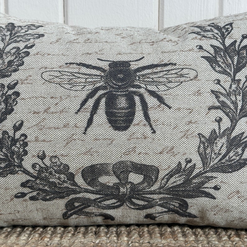 May include: A decorative pillow with a black and white design of a bee and floral wreath. The pillow is made of a light beige fabric with a subtle texture.