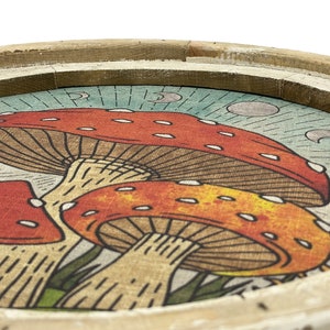 May include: A wooden tray with a circular design featuring three red and orange mushrooms with white spots on a blue and white background with crescent moons.