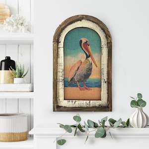 May include: A framed vintage-style print of a pelican standing on a beach with the ocean in the background. The frame is made of wood with a distressed white finish.