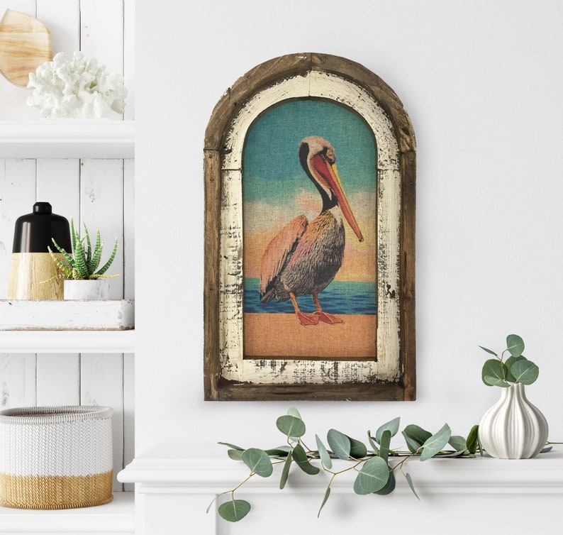 May include: A framed vintage-style print of a pelican standing on a beach with the ocean in the background. The frame is made of wood with a distressed white finish.