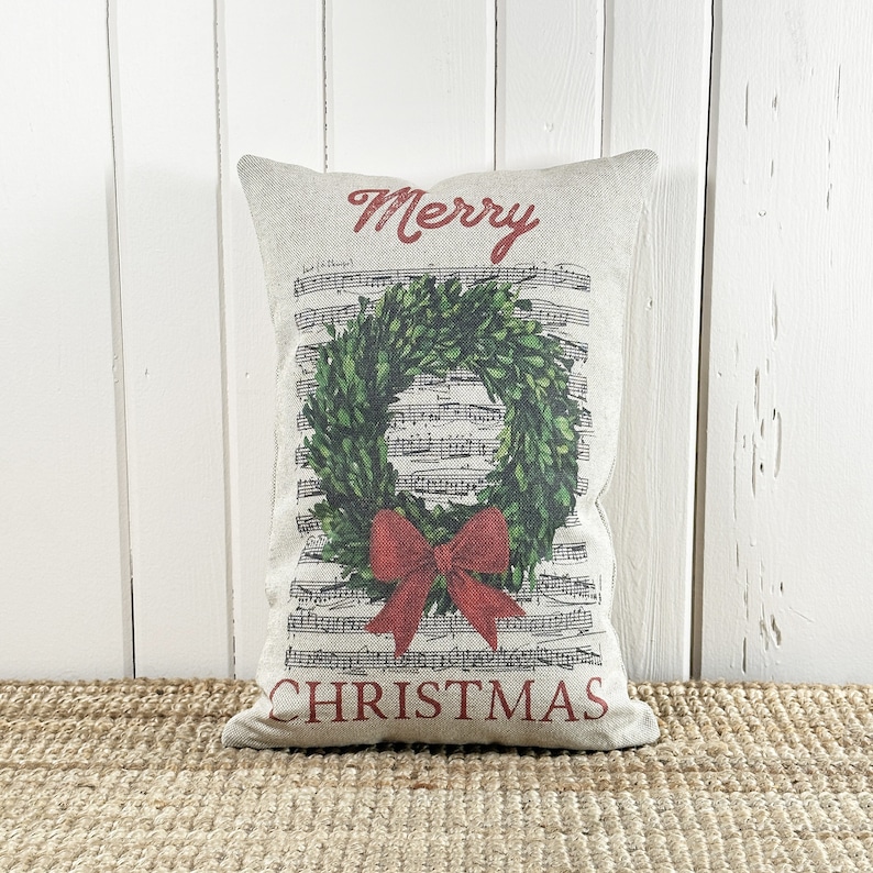 May include: A decorative throw pillow with a white background and a green holly wreath with a red bow. The pillow has the words "Merry Christmas" printed in red.