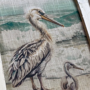 Pu&ograve; includere: Una stampa su tela beige di un pellicano e un uccello pi&ugrave; piccolo in piedi su una spiaggia. Il pellicano &egrave; marrone e bianco con un lungo becco. L'uccello pi&ugrave; piccolo &egrave; grigio e bianco. Lo sfondo &egrave; un'immagine sfocata di una spiaggia con onde e una montagna verde in lontananza.