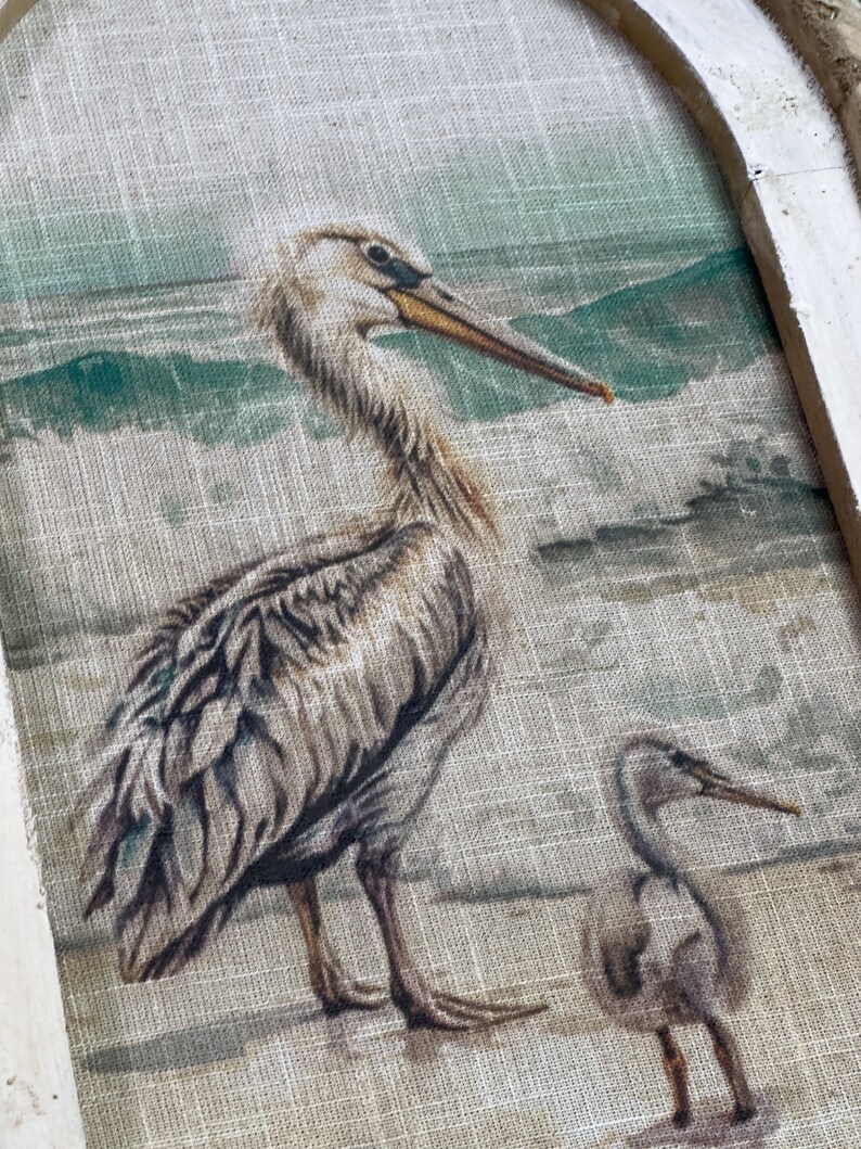 Pu&ograve; includere: Una stampa su tela beige di un pellicano e un uccello pi&ugrave; piccolo in piedi su una spiaggia. Il pellicano &egrave; marrone e bianco con un lungo becco. L'uccello pi&ugrave; piccolo &egrave; grigio e bianco. Lo sfondo &egrave; un'immagine sfocata di una spiaggia con onde e una montagna verde in lontananza.