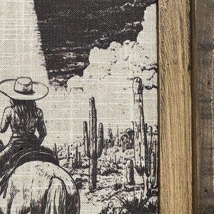 Puede incluir: Una ilustraci&oacute;n en blanco y negro de un vaquero montando a caballo a trav&eacute;s de un paisaje des&eacute;rtico con altos cactus. La imagen est&aacute; impresa en un lienzo y enmarcada con un marco de madera r&uacute;stico.