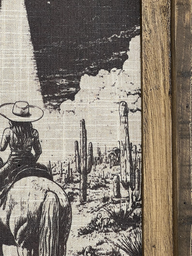 Puede incluir: Una ilustraci&oacute;n en blanco y negro de un vaquero montando a caballo a trav&eacute;s de un paisaje des&eacute;rtico con altos cactus. La imagen est&aacute; impresa en un lienzo y enmarcada con un marco de madera r&uacute;stico.