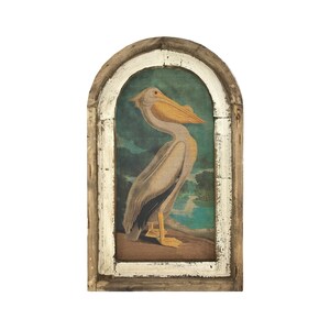 May include: A framed print of a pelican with a white body and yellow beak, standing on a brown surface with a blue and green background.