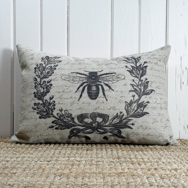 May include: A beige pillow with a black and white bee and floral wreath design. The pillow has a linen-like texture and a subtle script pattern.