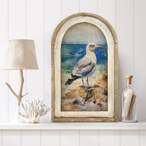 Op de afbeelding: Een ingelijst canvasprint van een meeuw die op een rots staat met de oceaan erachter. De lijst is van lichtbruin hout met een wit geschilderde boog.