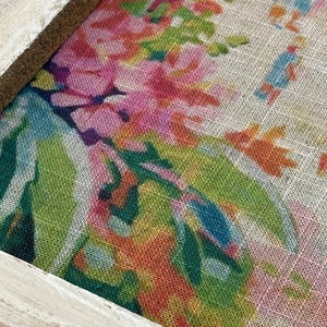 K&ouml;nnte beinhalten: Nahaufnahme eines gerahmten Stoffes mit einem Blumenmuster. Der Stoff ist hellbeige mit einer Vielzahl von rosa, orange, gr&uuml;nen und blauen Blumen. Der Rahmen ist hellbraun.