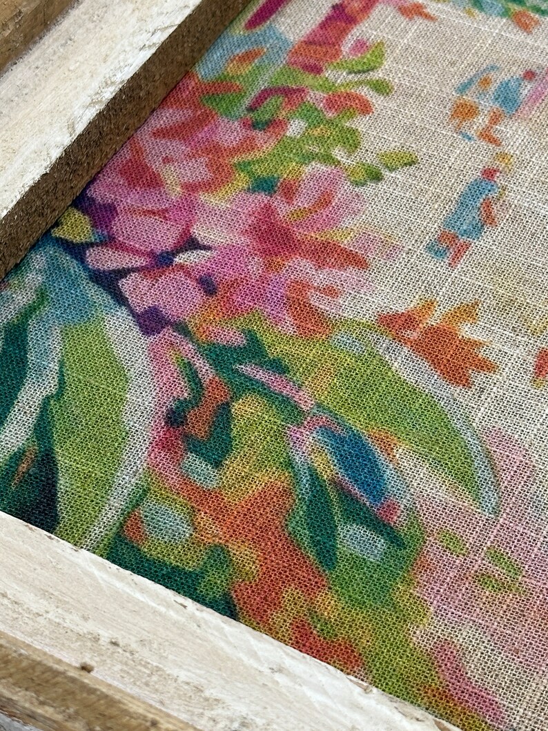 K&ouml;nnte beinhalten: Nahaufnahme eines gerahmten Stoffes mit einem Blumenmuster. Der Stoff ist hellbeige mit einer Vielzahl von rosa, orange, gr&uuml;nen und blauen Blumen. Der Rahmen ist hellbraun.