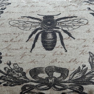 May include: A beige fabric pillow with a black and white bee design. The bee is centered on the pillow and has a detailed illustration of its wings and body. The background of the pillow is a light beige with a subtle texture.