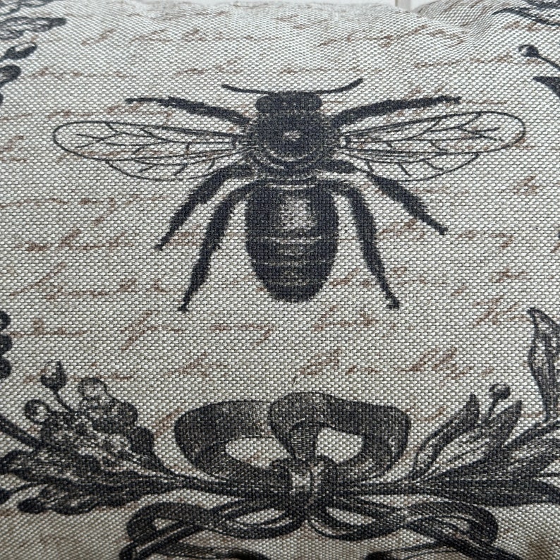 May include: A beige fabric pillow with a black and white bee design. The bee is centered on the pillow and has a detailed illustration of its wings and body. The background of the pillow is a light beige with a subtle texture.