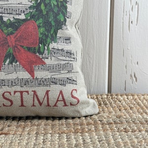 May include: A beige pillow with a red bow and a green wreath printed on it. The pillow has the word "STMAS" printed in red on the bottom.