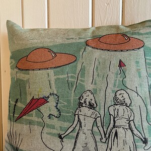 May include: A green and brown throw pillow with a design of two people walking away from two flying saucers. The people are wearing dresses and the saucers are orange and brown.