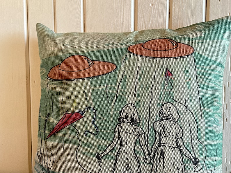 May include: A green and brown throw pillow with a design of two people walking away from two flying saucers. The people are wearing dresses and the saucers are orange and brown.
