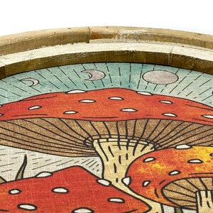 May include: A round wooden tray with a linen fabric insert featuring a whimsical illustration of red and orange mushrooms with white spots against a blue and green background. The mushrooms have brown stems and are surrounded by green grass.