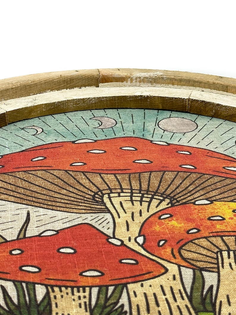 May include: A round wooden tray with a linen fabric insert featuring a whimsical illustration of red and orange mushrooms with white spots against a blue and green background. The mushrooms have brown stems and are surrounded by green grass.