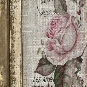 Pu&ograve; includere: Un'opera d'arte incorniciata con un'estetica vintage. L'immagine presenta una rosa rosa con foglie verdi su uno sfondo di lino. Il testo "Les Amateurs donnaient au Devant,  free me par" &egrave; stampato sul lino. La cornice &egrave; usurata e dipinta in un colore marrone chiaro.