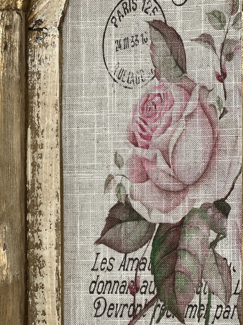 Pu&ograve; includere: Un'opera d'arte incorniciata con un'estetica vintage. L'immagine presenta una rosa rosa con foglie verdi su uno sfondo di lino. Il testo "Les Amateurs donnaient au Devant,  free me par" &egrave; stampato sul lino. La cornice &egrave; usurata e dipinta in un colore marrone chiaro.