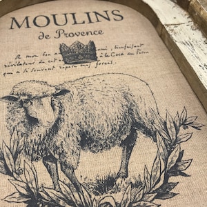 Op de afbeelding: Een rustieke houten dienblad met een witgekalkte afwerking heeft een gedrukt ontwerp van een schaap met een kroon erboven. De tekst "MOULINS de Provence" is boven het schaap gedrukt. De tekst onder het schaap luidt: "A mon bon z&eacute;v&eacute;lateur de cat Coca du P&eacute;ron qui a Si Souvant r&eacute;pare mas foreas."