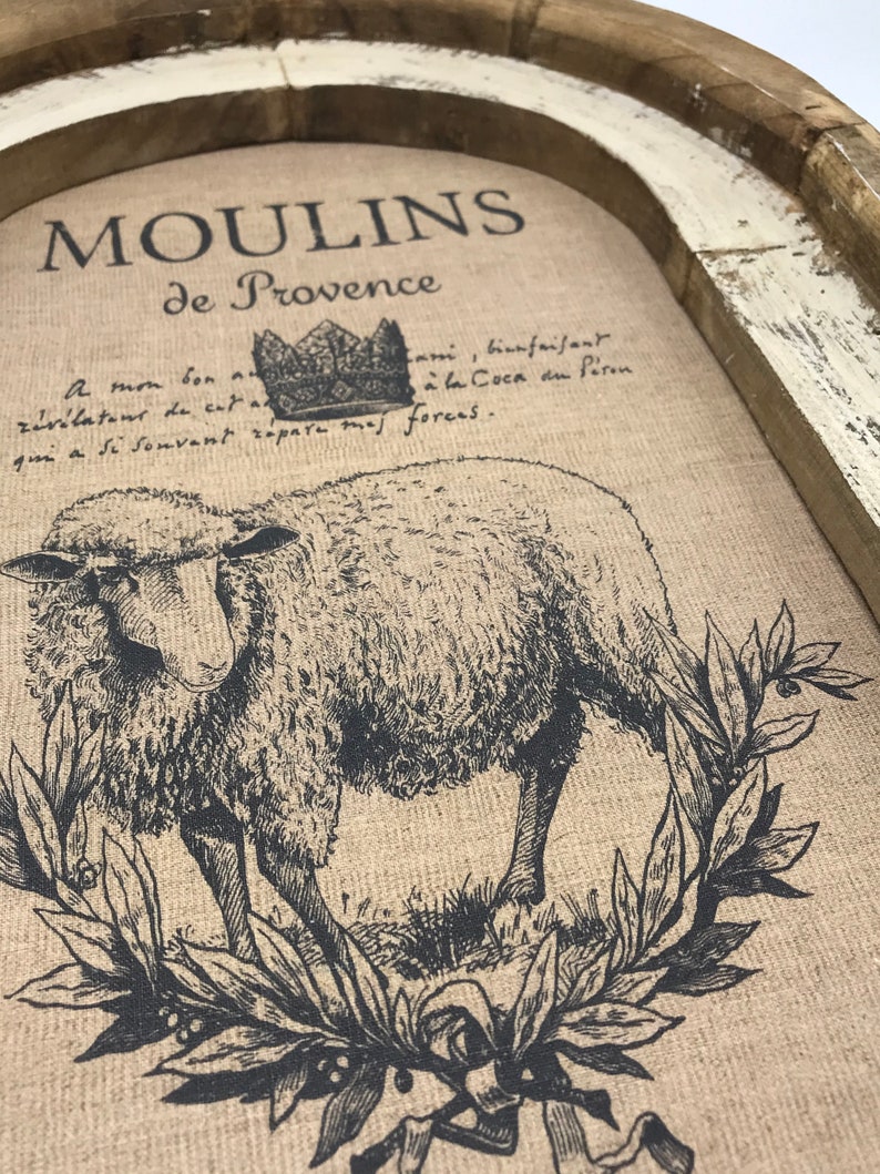 Op de afbeelding: Een rustieke houten dienblad met een witgekalkte afwerking heeft een gedrukt ontwerp van een schaap met een kroon erboven. De tekst "MOULINS de Provence" is boven het schaap gedrukt. De tekst onder het schaap luidt: "A mon bon z&eacute;v&eacute;lateur de cat Coca du P&eacute;ron qui a Si Souvant r&eacute;pare mas foreas."