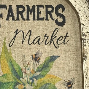 May include: A rustic wooden sign with a distressed white paint finish. The sign features the text "FARMERS Market" in black lettering and a painted image of a lemon tree with white blossoms and two bumblebees.